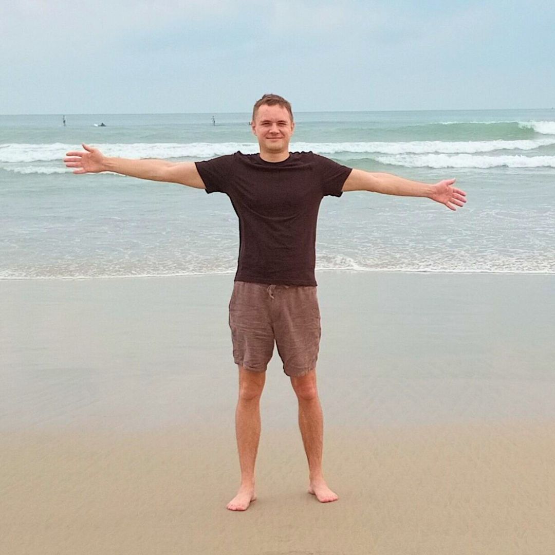 Marcus King Wolff standing barefoot on the beach with arms outstretched, facing the camera, with ocean waves in the background.